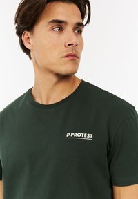 Jonge man met bruin haar die opzij kijkt, gekleed in een donkergroen T-shirt met het woord "PROTEST" in het wit op de borst gedrukt.