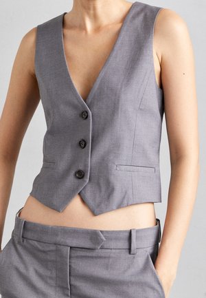 Personne portant un gilet gris sans manches boutonné et un pantalon gris assorti, les mains dans les poches, sur un fond uni.