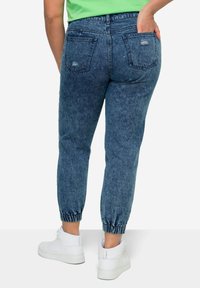 Joggers de mezclilla en azul medio con puños elásticos en los tobillos. Presenta detalles desgastados y bolsillos traseros tradicionales. Combinados con zapatillas blancas.