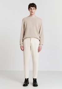 Junger Mann, der einen beigen Pullover, eine cremefarbene Hose und schwarze Stiefel trägt, steht vor einem schlichten hellen Hintergrund.