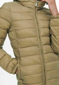 Persona que lleva una chaqueta acolchada de color verde oliva con cremallera en el frente, con una mano en el bolsillo y la otra ajustando el cuello.