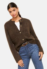 Brūns adīts kardigāns ar apaļu kakla izgriezumu, pogu aizdari, divām priekšējām kabatām un ribētām piedurknēm. Saskaņots ar ziliem džinsiem un brūnu jostu.