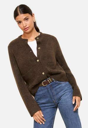 Brauner Strickcardigan mit Rundhalsausschnitt, Knopfverschluss, zwei Fronttaschen und gerippten Bündchen. Kombiniert mit blauen Jeans und einem braunen Gürtel.