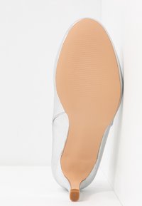 Semelle d'une chaussure à talon haut argentée avec une semelle en caoutchouc beige et un petit embout de talon, montrée droite sur fond blanc.