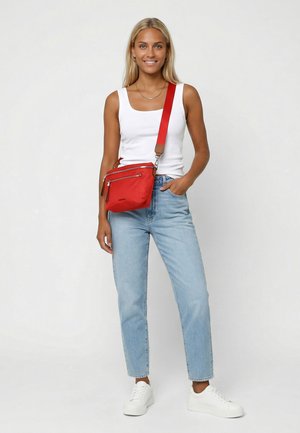 Ung kvinde iført hvid tanktop, lyseblå jeans, hvide sneakers og en rød skuldertaske, stående mod en ensfarvet lys baggrund.