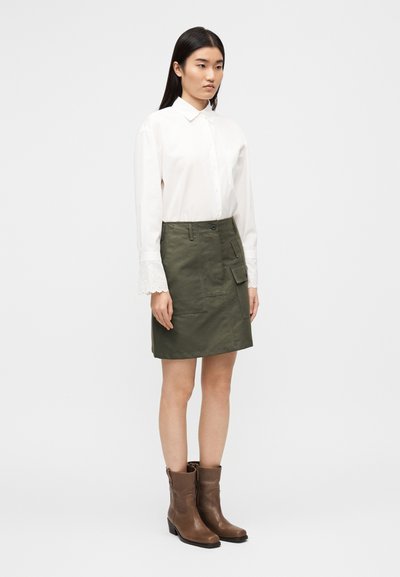 Λευκό πουκάμισο με κουμπιά και δαντελένια μανσέτα, φούστα ελιάς με πλαϊνές τσέπες και καφέ μποτάκια στον αστράγαλο με χαμηλό τακούνι.