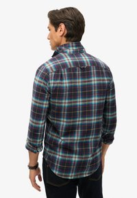 Camicia di flanella a quadri nei colori blu navy, verde e bordeaux con maniche arrotolate. Presenta un colletto button-down e uno sprone posteriore per la struttura.