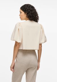 Beige kurzes Oberteil mit Puffärmeln und glatter Textur. Kombiniert mit hellbeigen, strukturierten, hochgeschnittenen Hosen. Sauberes, minimalistisches Design.
