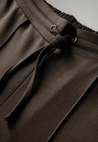 Bruine tailleband met trekkoord en een gladde textuur, voorzien van geplooide details. Het materiaal is zacht en rekbaar, wat zorgt voor een comfortabele pasvorm.