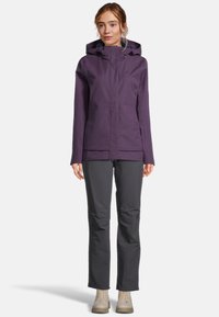 Giacca impermeabile viola con cappuccio, due tasche frontali e dettagli in zip, abbinata a pantaloni grigi e stivali beige stringati.