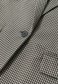 Czarny i kremowy materiał w wzór pepitki o gładkiej fakturze, z czarnym guzikiem i ozdobą w postaci kieszeni naszywanej.