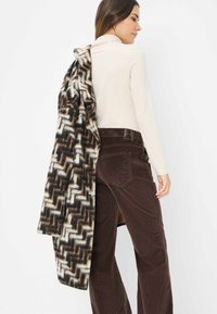 Bruine corduroy broek met een hoge taille, gecombineerd met een crèmekleurige coltrui en een patroonjas met zwart, wit en tan zigzagpatronen.