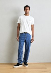 Camiseta blanca de manga corta con un pequeño logo, combinada con jeans azules de corte holgado y zapatillas negras con suelas blancas sobre un suelo de madera.