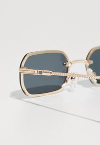 Óculos de sol quadrados com uma armação de metal dourado, lentes escuras e hastes com textura. Possui Almofadas ajustáveis no nariz para conforto.