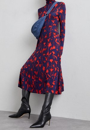 Kvinde iført en navyblå kjole med rød blomsterprint, sorte spidse hæle støvler og en blå crossbody-taske foran en ensfarvet grå væg.