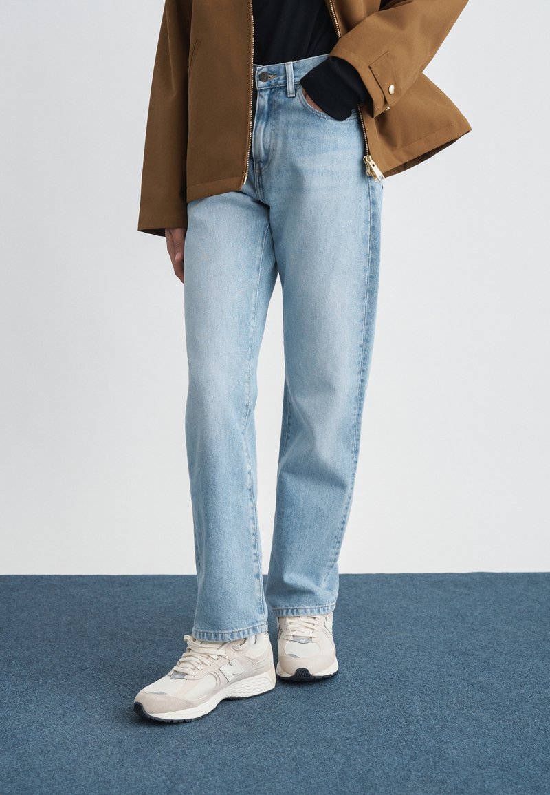 Persoon die lichtblauwe jeans met rechte pijpen draagt, beige sneakers, een zwart shirt en een bruine jas met hand in de zak, staand op een blauw tapijt.