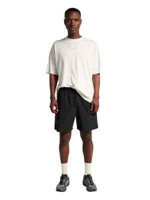 Mann steht und trägt ein weißes oversized T-Shirt, schwarze Shorts, weiße Socken und graue Sportschuhe vor weißem Hintergrund.