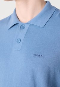 Polo bleu clair avec deux boutons et logo brodé "BOSS" sur la poitrine gauche, porté par une personne avec un cou et une mâchoire visibles.