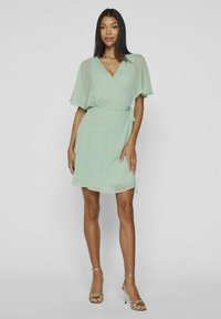 Zelené oblečenie vo forme šiat s krátkymi, voľnými rukávmi. Ľahká, textúrovaná látka s výstrihom do V a detailom na bočnom zaviazaní. V kombinácii s metalicky lesklými opätkami.