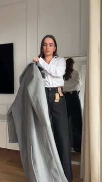 Cappotto grigio dalla texture morbida, tenuto da una persona con una camicia bianca e jeans neri. Le etichette attaccate al cappotto mostrano i dettagli del prezzo.