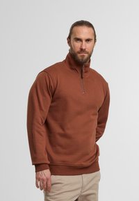 Brun zip-up sweatshirt i blødt stof, med høj krave og ribbede manchetter og kanten, båret med lyse bukser.