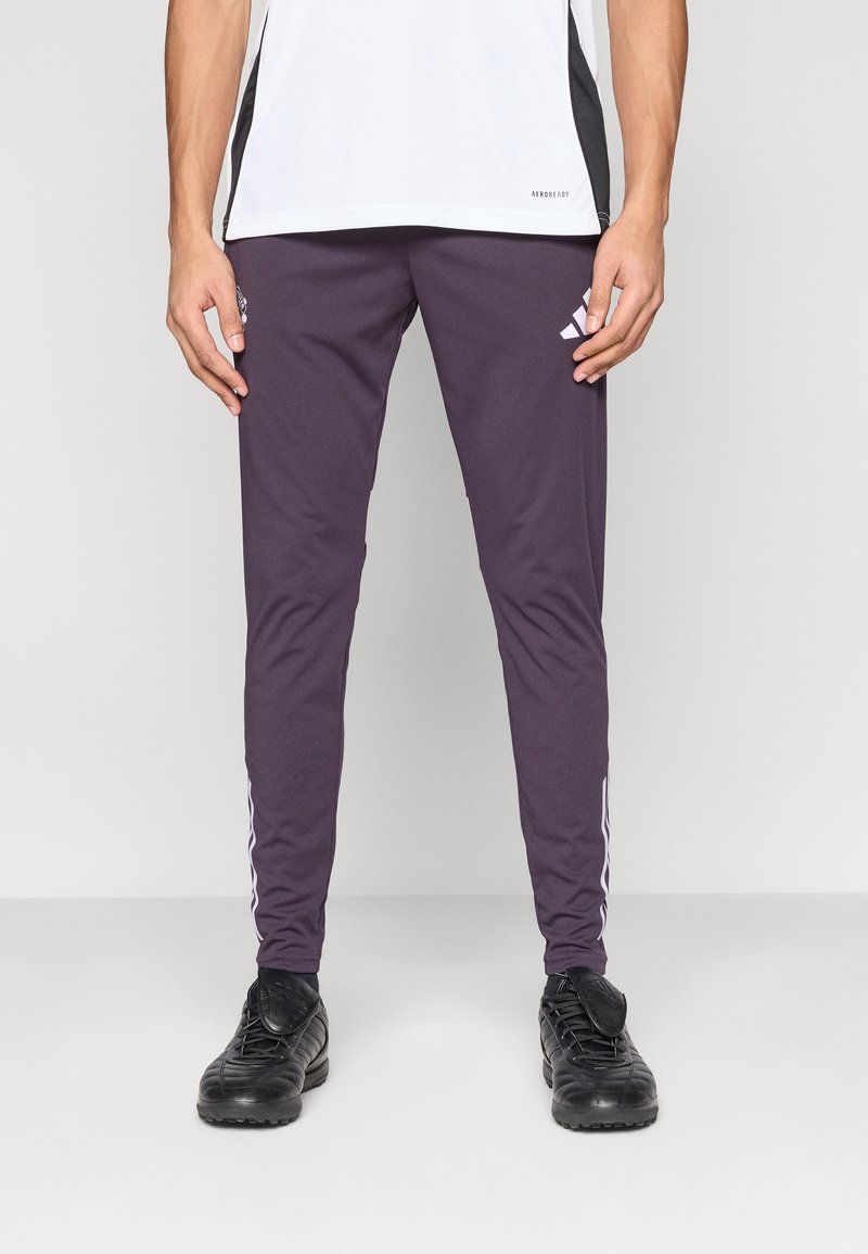 Pantalones deportivos para hombre en morado oscuro, corte ajustado. Hechos de tela ligera. Presentan rayas laterales blancas y logo, con zapatillas deportivas negras.