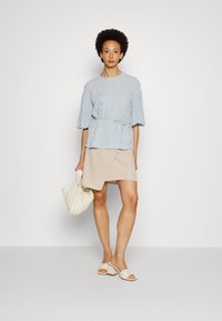 Blusa texturada azul clara com mangas largas e cintura ajustável por cordão, combinada com uma saia envolvente bege e sandálias tipo slide em branco sujo. Inclui uma bolsa bege.