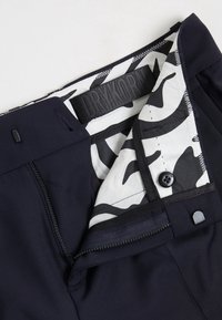 Granatowe spodnie z czarnymi elementami, z białą podszewką w kamuflażowy wzór w obrębie pasa i strefy zamka błyskawicznego. Widoczna etykieta marki.