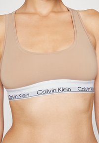 Sostenitore sportivo beige realizzato in tessuto morbido. Presenta una larga fascia bianca con il logo nero "Calvin Klein". Texture liscia e design semplice.