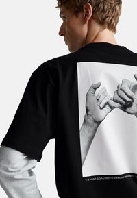 Mladý muž má na sobě černé tričko s černobílou fotografií dvou rukou, které se propojují malíčky, na zadní straně, pod kterým má šedý dlouhý rukáv.