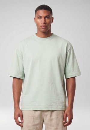 Jeune homme tourné vers l'avant, portant un t-shirt oversize vert clair à manches courtes et un pantalon beige sur un fond uni.