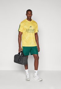 Κίτρινη αθλητική μπλούζα Adidas με πράσινες λεπτομέρειες και λογότυπο, συνδυασμένη με πράσινες αθλητικές βερμούδες. Κρατώντας μια μαύρη τσάντα γυμναστηρίου, φορώντας άσπρες κάλτσες και παπούτσια.