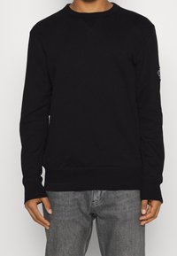 Felpa nera con colletto rotondo, polsini e orlo a coste. Tessuto morbido, con una patch logo sulla manica sinistra. Modello che indossa jeans grigi.