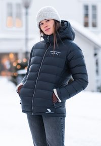 Wyściełana czarna kurtka z kapturem, z logo na górnej lewej części, teksturowanymi mankietami i kieszeniami zapinanymi na zamek, w połączeniu z szarymi dżinsami i dzianą czapką.