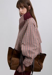 Rutig oversized jacka i rött och vitt med en hög vinröd krage, kombinerad med en brun mockatoteväska med mörka handtag.