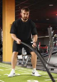 Zwart shirt met korte mouwen en shorts gedragen door een persoon die een battle rope gebruikt op een groene gymvloer, met witte sneakers en fitnessapparatuur op de achtergrond.