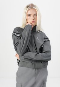 Veste grise zippée avec manches longues, dotée de rayures blanches sur les épaules et d'un petit logo ; texture lisse et design court.
