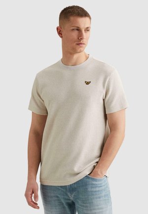 Muž nosiaci béžovú textúrovanú košeľu s krátkym rukávom a malým žlto-čiernym logom vtáka na hrudi, modré džínsy, stojaci s jednou rukou v vrecku.