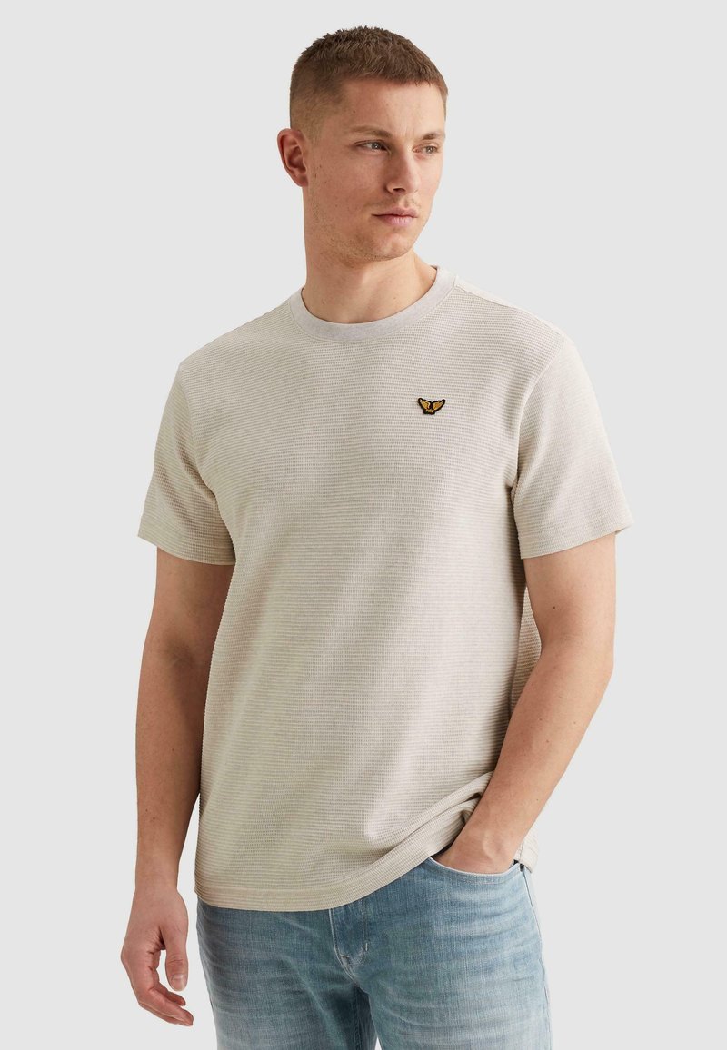 Homme portant une chemise texturée beige à manches courtes avec un petit logo d'oiseau jaune et noir sur la poitrine, jean bleu, debout avec une main dans la poche.