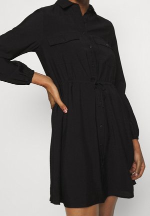 Kobieta w czarnej, zapinanej na guziki sukience z długim rękawem, z kieszeniami na piersi i sznurkiem w talii, ręka oparta na biodrze.