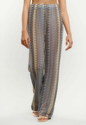 Széles szárú nadrágok függőleges, hullámos csíkokkal kék, bézs, fekete és fehér színekben, amelyeket egy fehér háttér előtt álló, szandált viselő személy visel.