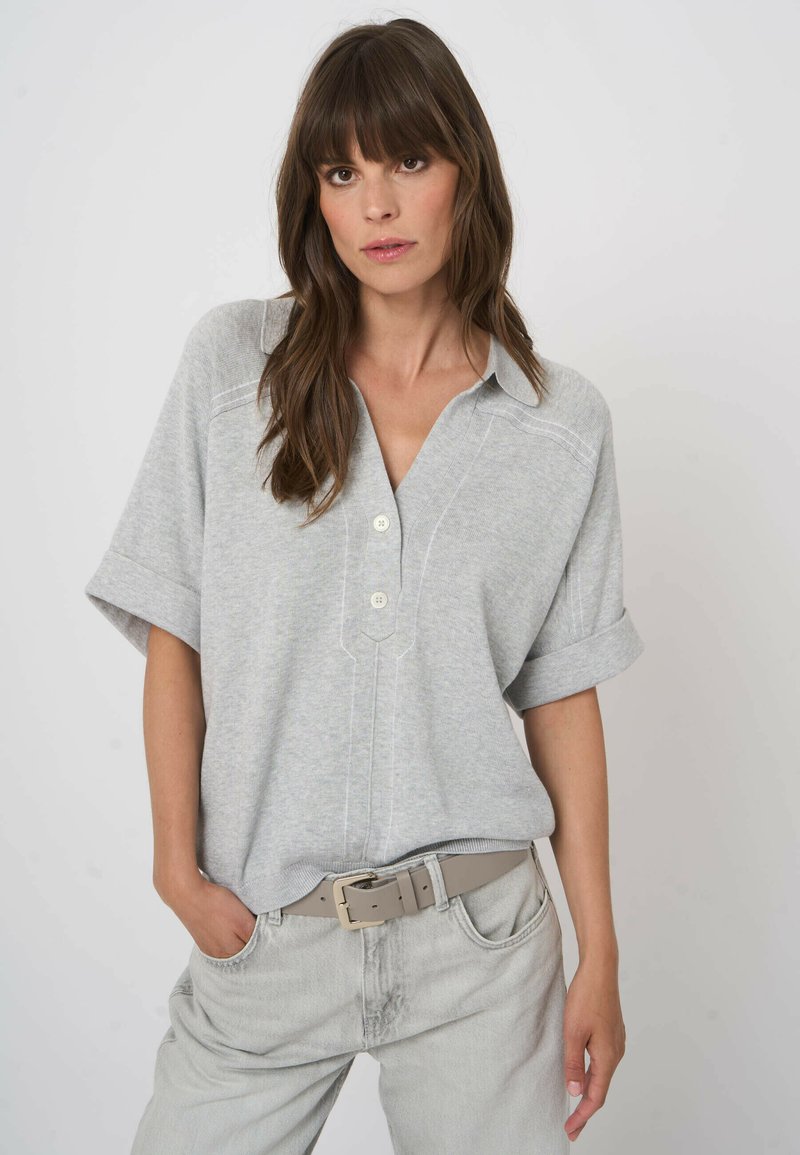 Frau mit langen braunen Haaren trägt ein hellgraues kurzärmeliges Hemd mit Knöpfen, das in eine hellgraue Jeans mit passendem Gürtel gesteckt ist.