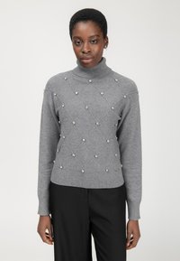 Maglione grigio lavorato a maglia con collo alto, motivo a rombi e decorazioni di perle; polsini e orlo a costine. Pantaloni neri sotto.