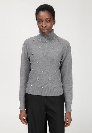 Maglione grigio lavorato a maglia con collo alto, motivo a rombi e decorazioni di perle; polsini e orlo a costine. Pantaloni neri sotto.
