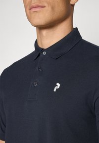 Námornícké polo tričko vyrobené z textúrovaného materiálu, má tri gombíky v oblasti krku a strieborný logový akcent na ľavej hrudi.