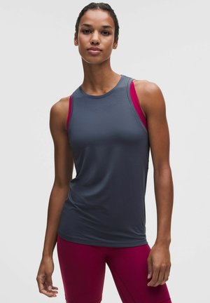 Frau mit ärmellosem dunkelgrauem Sporttop über einem magentafarbenen Sport-BH und passenden magentafarbenen Leggings steht vor einem hellgrauen Hintergrund.