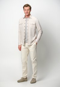 Camicia button-up beige chiaro con due tasche sul petto sopra una maglietta grigia a girocollo, abbinata a pantaloni bianchi e sneakers beige.