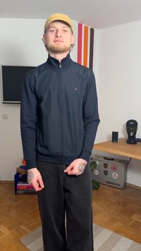 Navy zip-up jacket with high collar, ribbed cuffs, and small logo detail. Paired with loose black pants. Background features a wooden table.