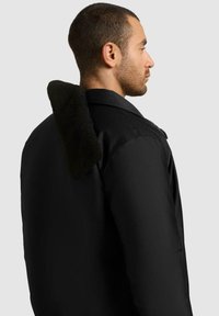 Svart jakke med en strukturert, mørkegrønn pelskrage. Har en glatt finish og strukturerte skuldre, vist fra siden.