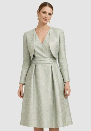 Donna che indossa un abito jacquard floreale verde chiaro con vita plissettata e giacca corta abbinata, accessoriata con orecchini pendenti.